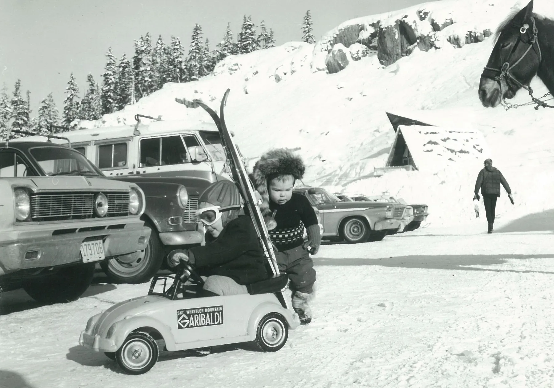 Black-and-white photo of a child in a winter hat holding skis beside vintage cars in the snow, early 1970s